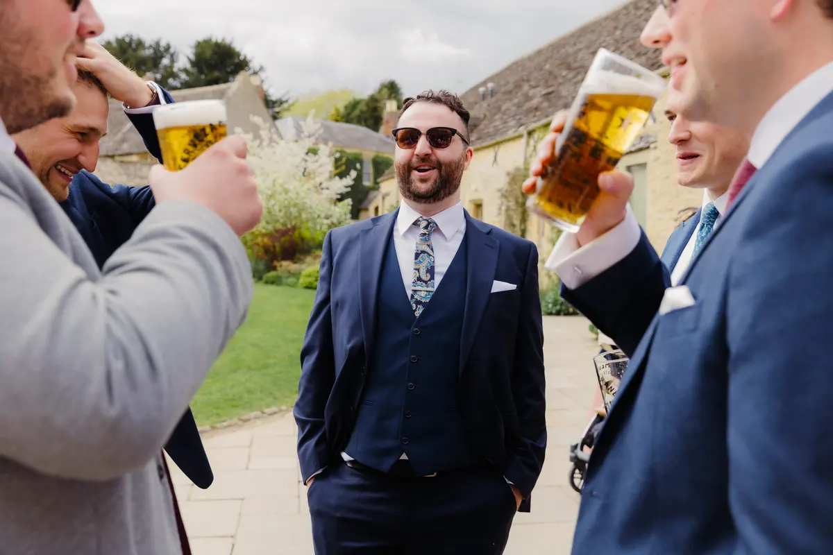 Groomsmen in navy suits drinking pints of beer and laughing in the courtyard at Caswell House