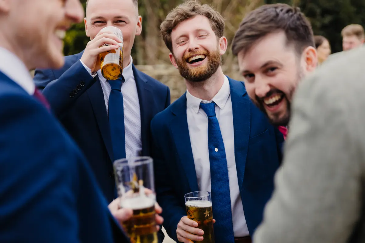Wedding guests in blue suits laughing together over pints of beer at Caswell House