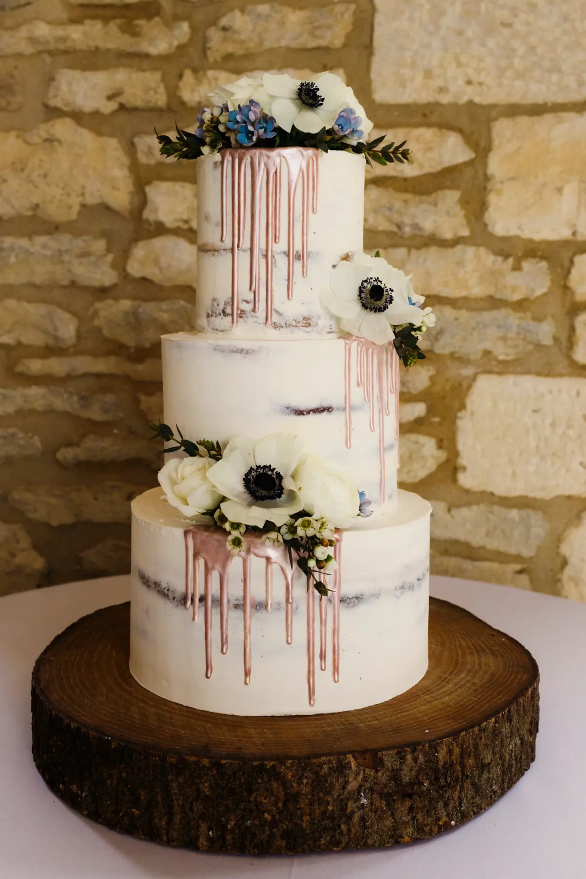 Three-tier semi-naked wedding cake with pink drip and white anemones on a log slice at Caswell House