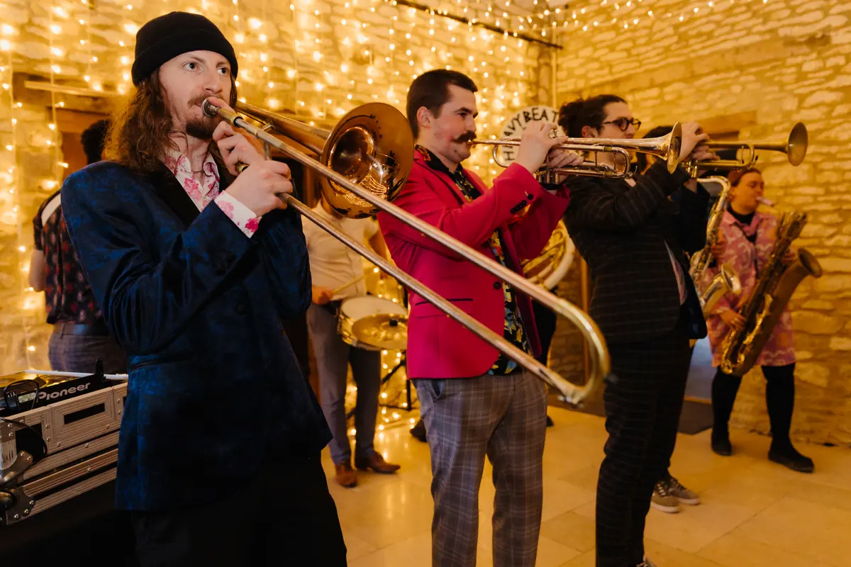 Brass band playing trombone, trumpets and saxophone against a stone wall lit with fairy lights