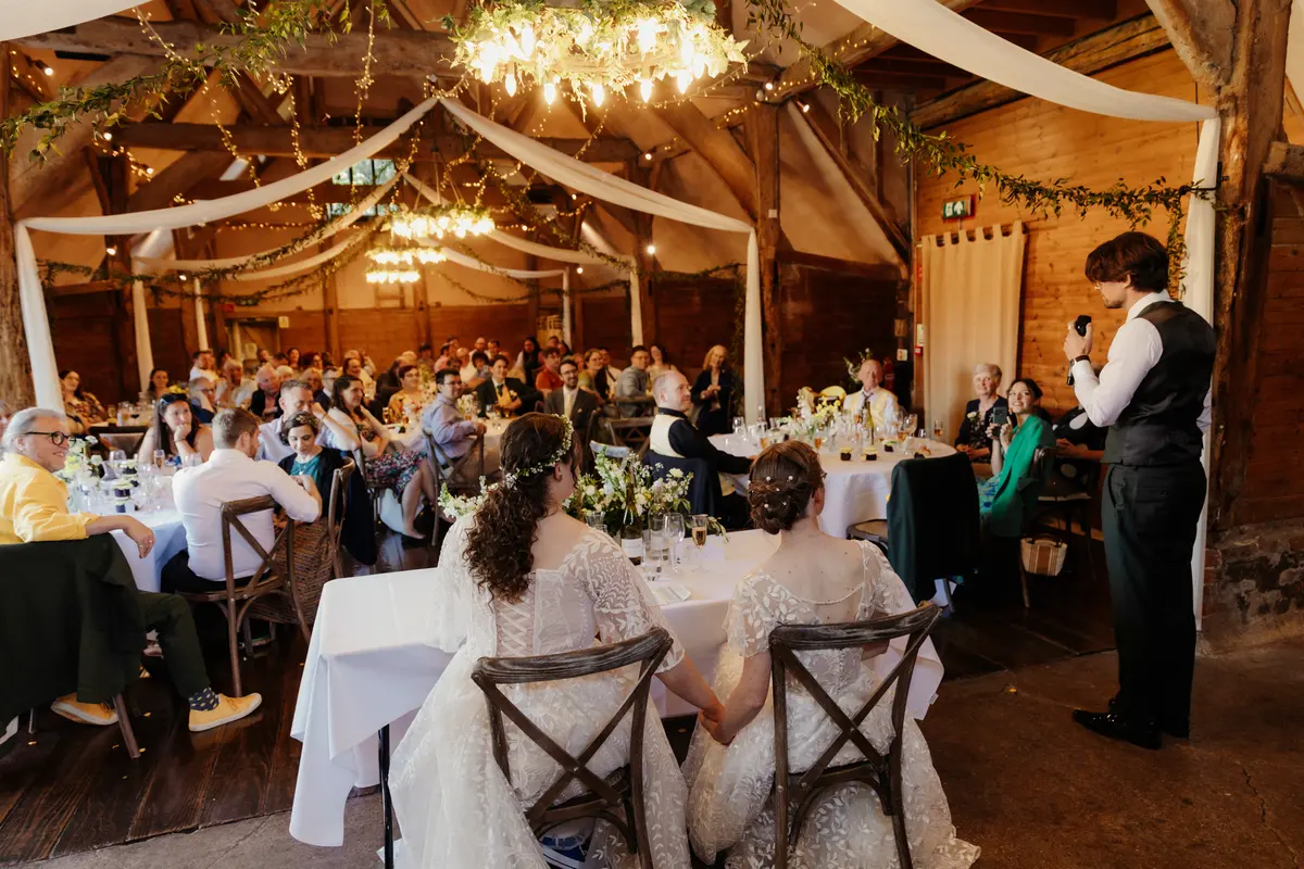 Two brides holding hands at their table as a guest gives a speech at Lains Barn
