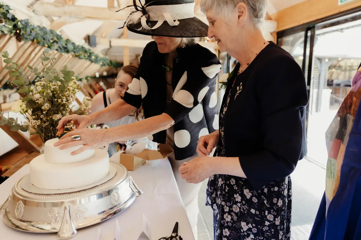 Two older women cutting a white wedding cake on a silver stand at Lains Barn