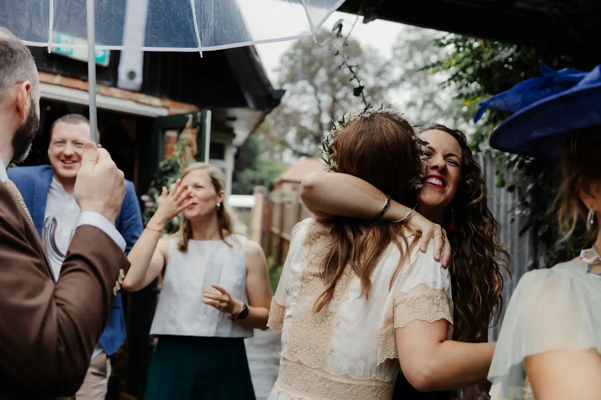 Bride hugging a smiling guest under a clear umbrella on a rainy day