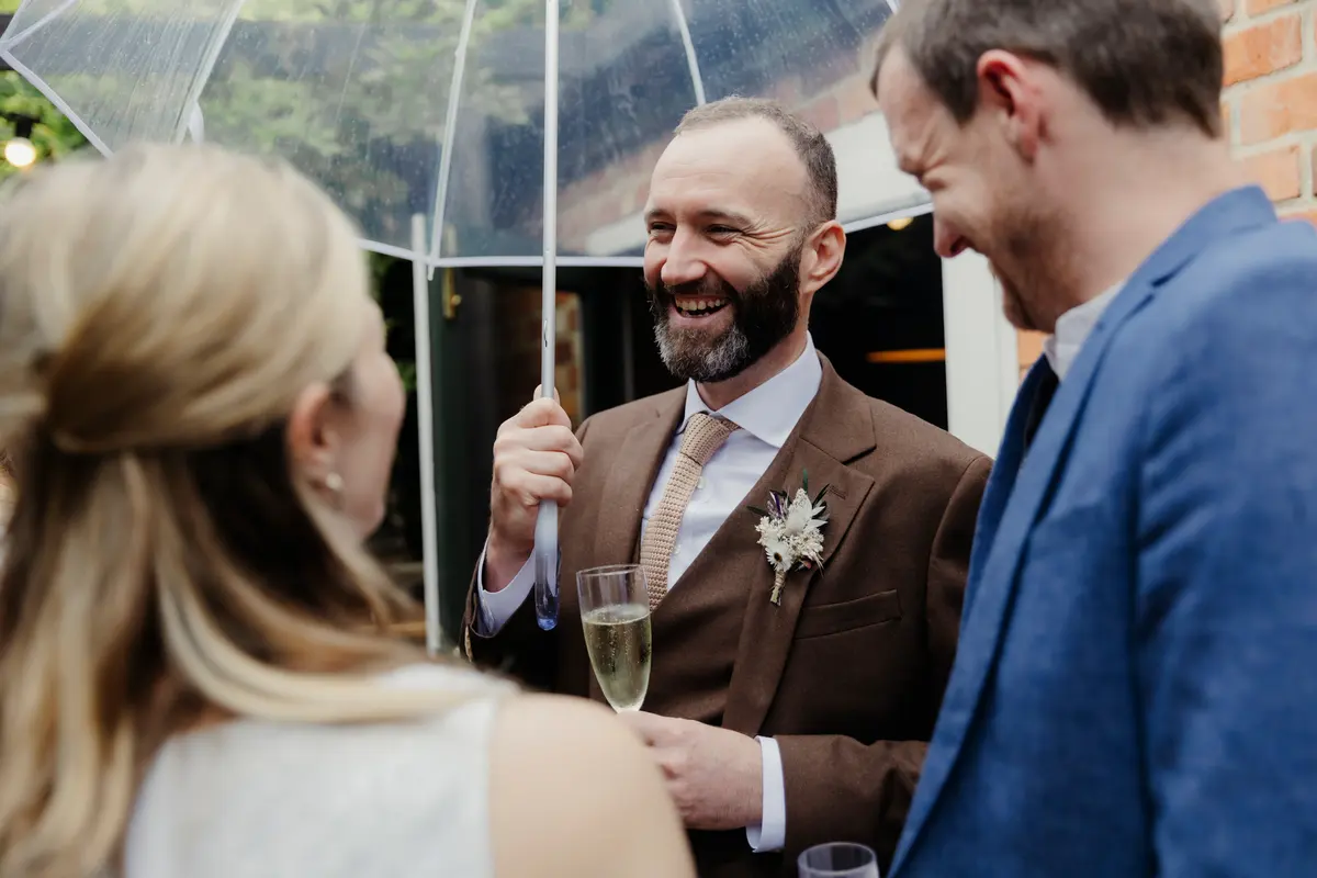Groom laughing under a clear umbrella with a glass of fizz, chatting with guests
