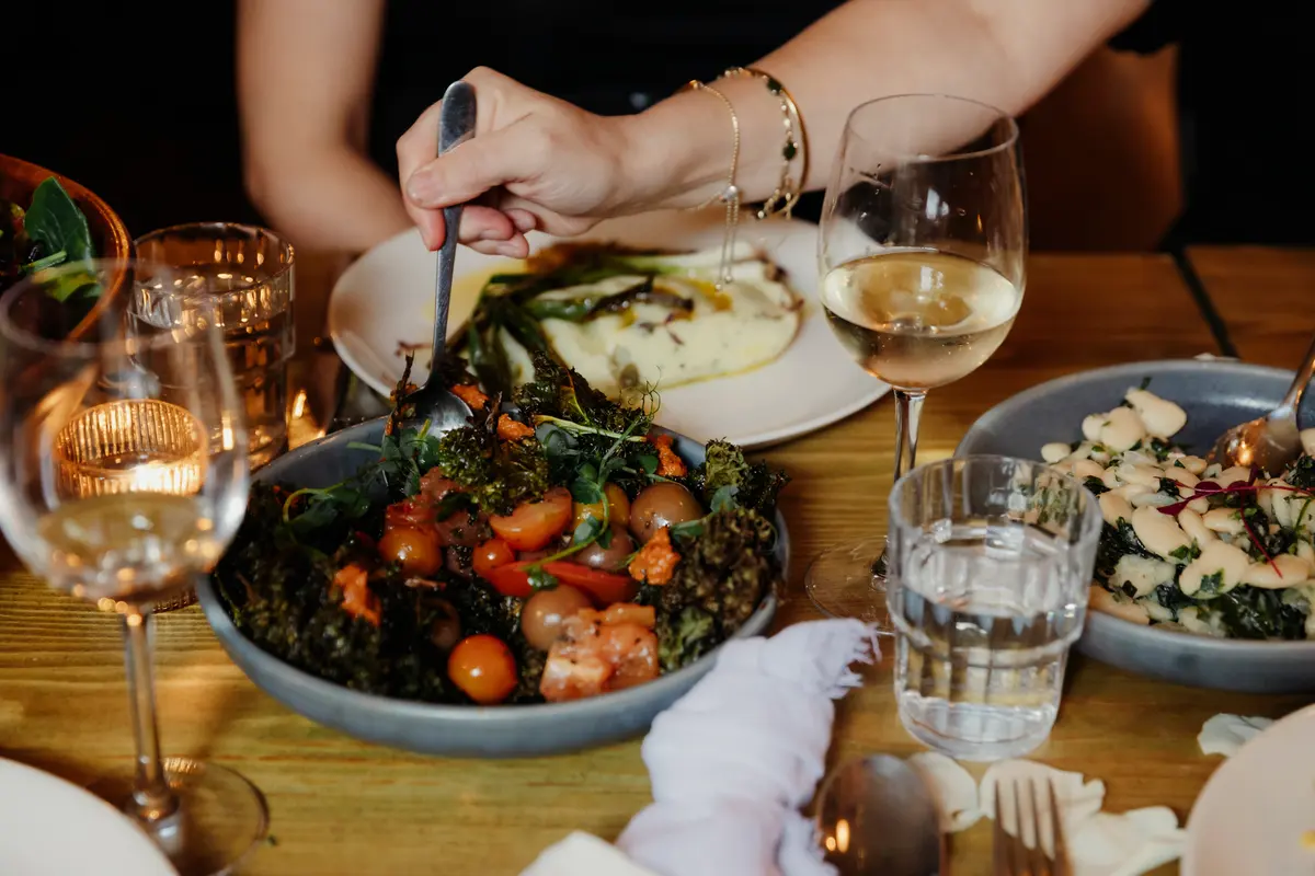 Guest spooning from a bowl of roasted tomatoes and kale beside glasses of white wine
