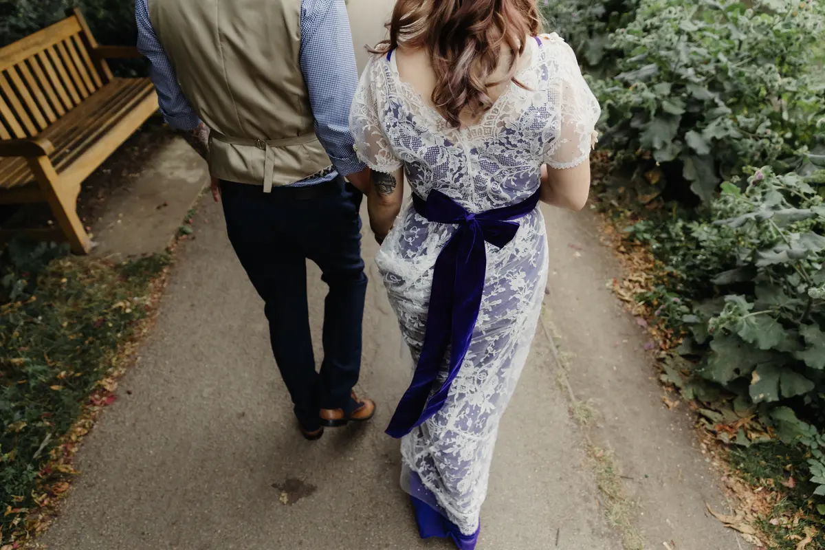 Bride in a lace dress with a purple sash walking hand in hand with the groom along a garden path