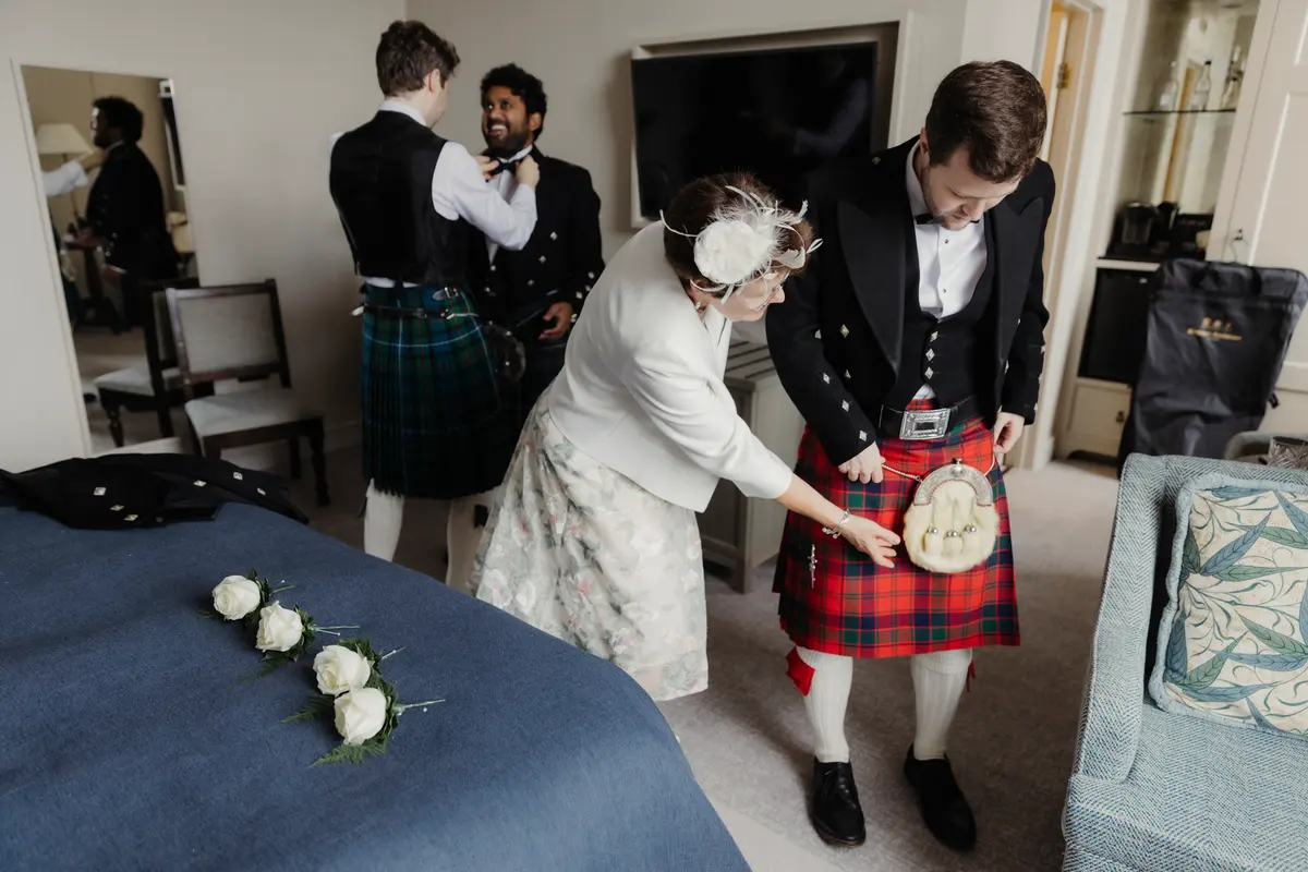 Woman adjusting a groom's sporran as men in kilts get ready in a hotel room