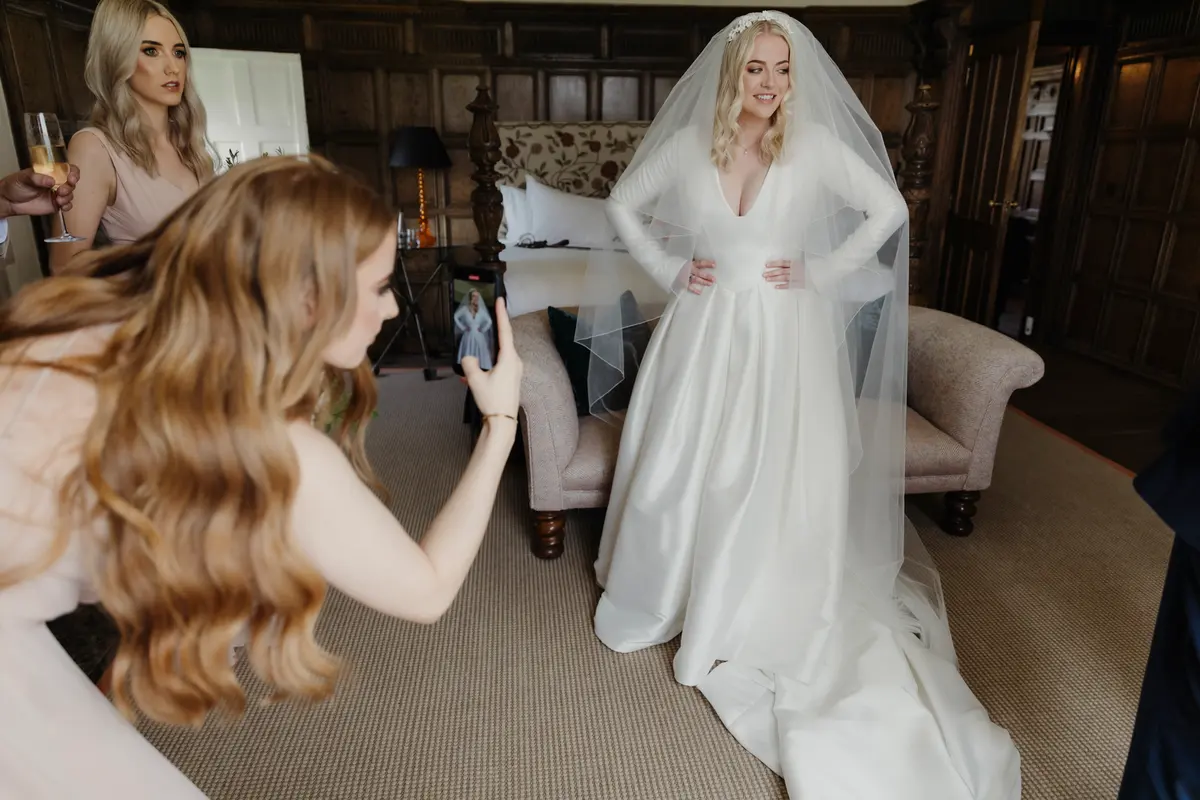 Bridesmaid photographing the bride on her phone in a wood-panelled room as the bride poses in her long veil