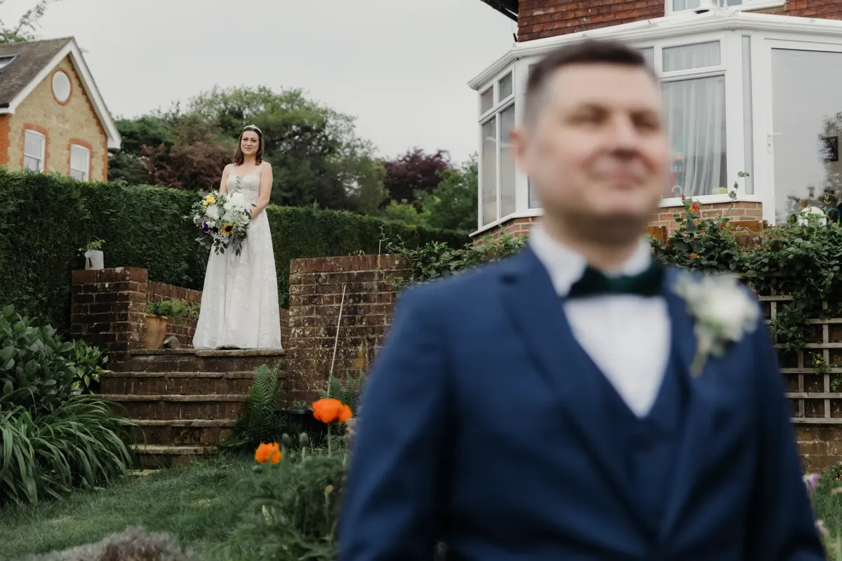 Bride waiting at the top of garden steps as the groom stands in the foreground for a first look