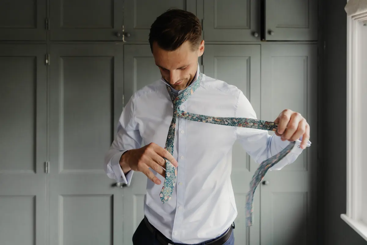 Groom in a white shirt tying a floral tie in front of grey panelled cupboards