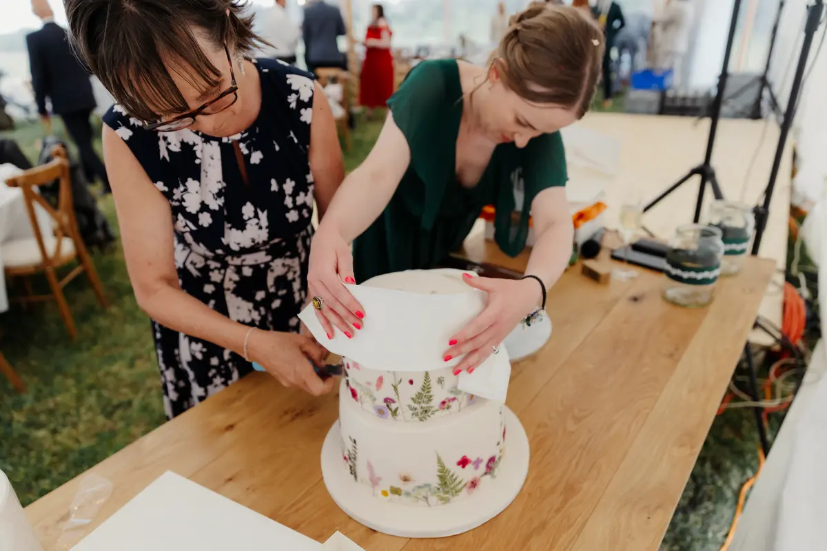 Two women placing a tier on a wedding cake decorated with pressed flowers in a marquee