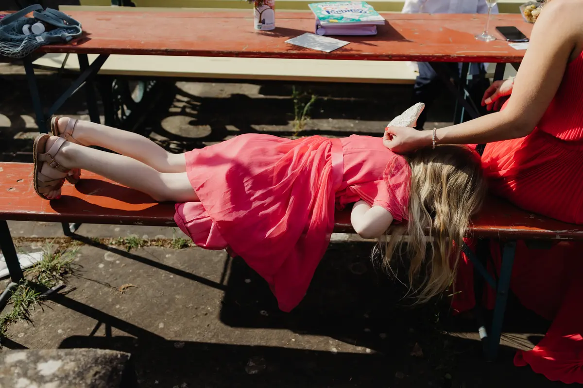 Girl in a pink dress lying face down on a bench as a woman strokes her hair at a sunny outdoor wedding